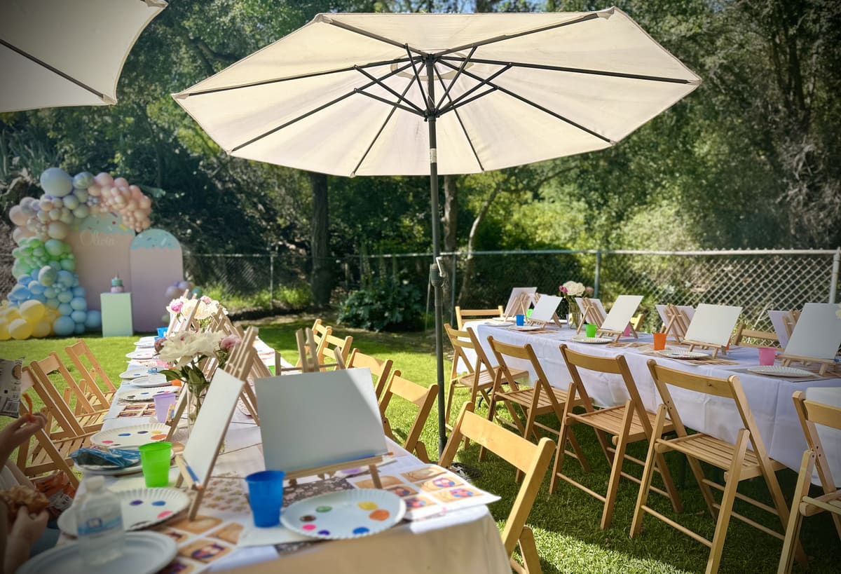A backyard set for a celebration: long tables of easels, a shade umbrella and a pastel balloon arch